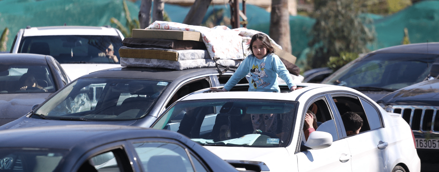A young girl stands inside a car with her upper body through the sunroof as vehicles queue in traffic in Lebanon during mass displacement.
